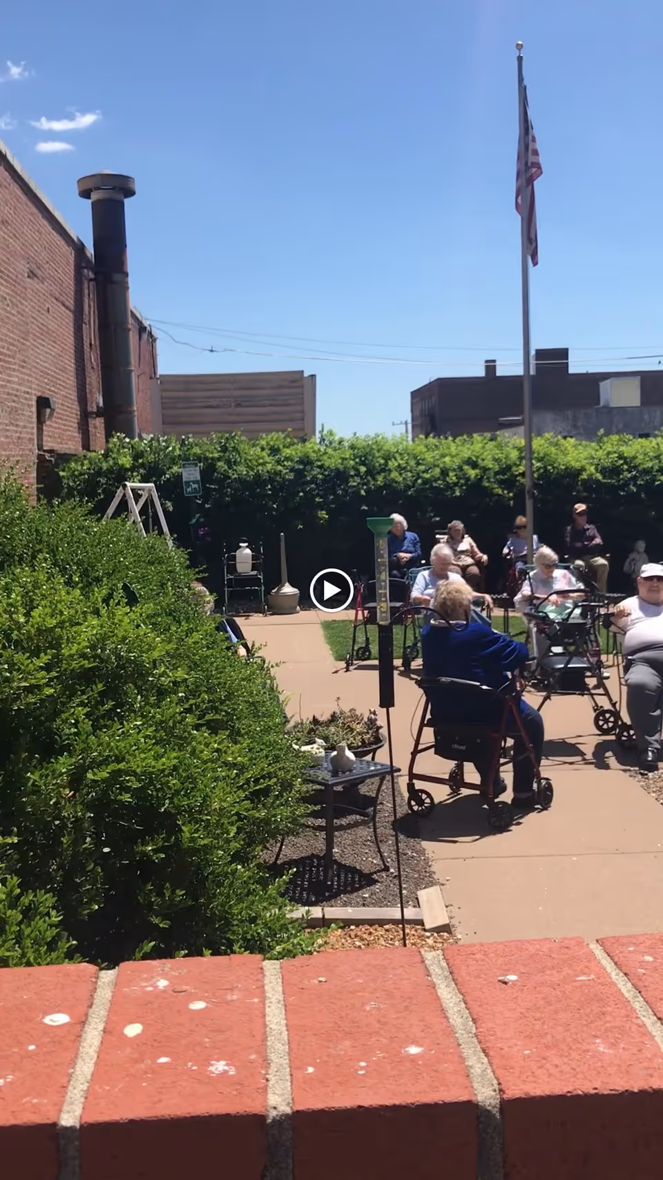 Outdoor patio area with several elderly people sitting on chairs and using walkers, surrounded by green bushes and a brick wall in the foreground. An American flag is visible on a flagpole against a clear blue sky.