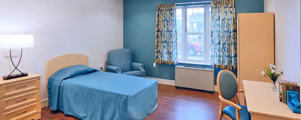 A senior living bedroom featuring a single bed with a blue bedspread, a wooden nightstand with a lamp, a comfortable armchair, a window with patterned curtains, a wooden wardrobe, and a desk with a chair and decorative items.