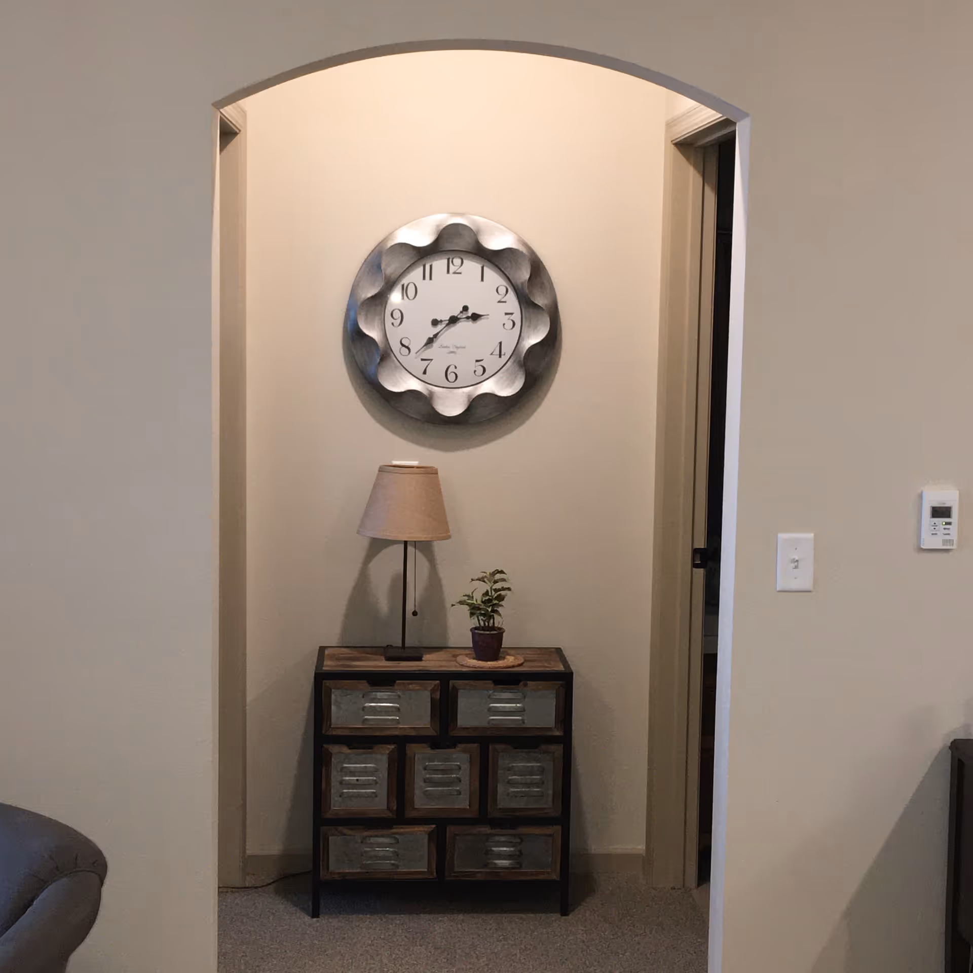 A small hallway area with an arched entryway. On the wall is a decorative round clock with a wavy metallic frame. Below the clock is a small cabinet with nine metal drawers, on top of which sits a table lamp with a beige shade and a small potted plant. To the right of the archway is a light switch and a thermostat on the wall. Part of a gray upholstered chair is visible on the left side of the image.