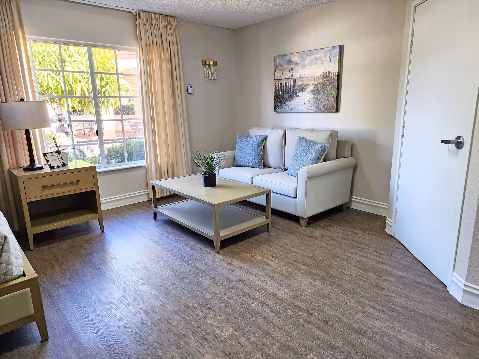 A cozy living room area with a white loveseat adorned with two blue cushions, a light wooden coffee table with a small potted plant, a side table with a lamp and a framed photo, beige curtains covering a window, and a wall painting depicting a beach scene.