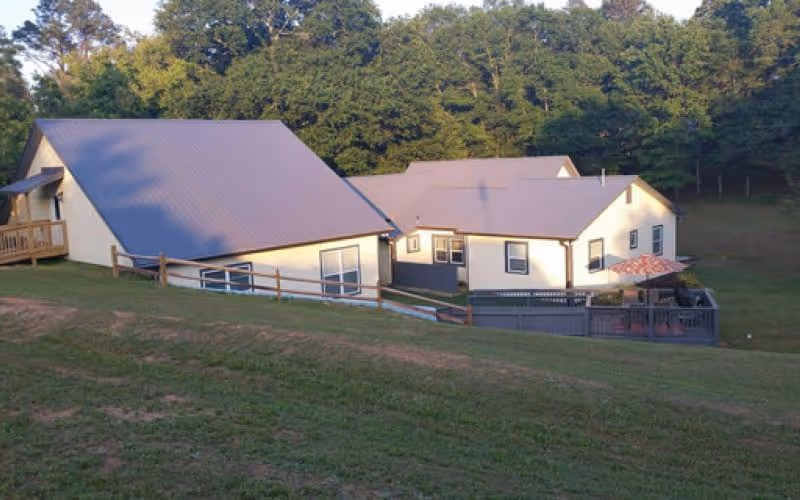 Exterior view of a single-story building with light-colored walls and a metal roof, situated on a grassy hill with a wooden fence and surrounded by trees in the background.