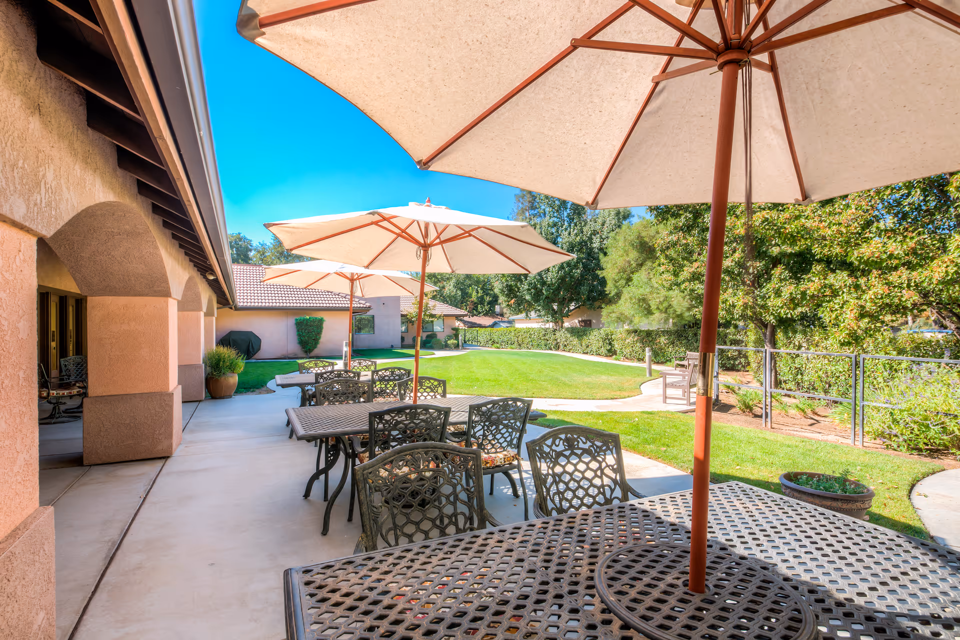 Outdoor patio area at Summerfield Memory Care Of Fresno with metal tables and chairs under large beige umbrellas, adjacent to a building with arched doorways, overlooking a well-maintained lawn and trees under a clear blue sky.