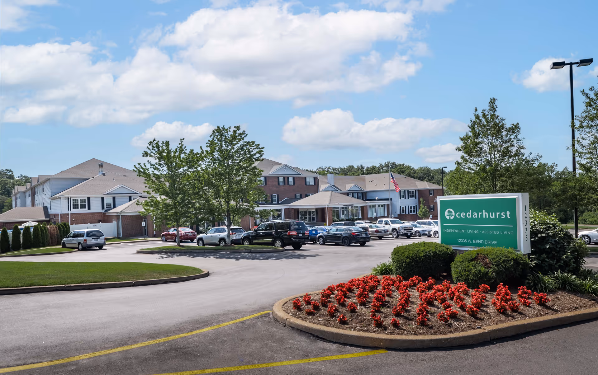 Exterior view of Cedarhurst Senior Living of Tesson Heights facility with a parking lot, several cars, trees, and a landscaped flower bed with red flowers in front of a green sign displaying the facility name and address.
