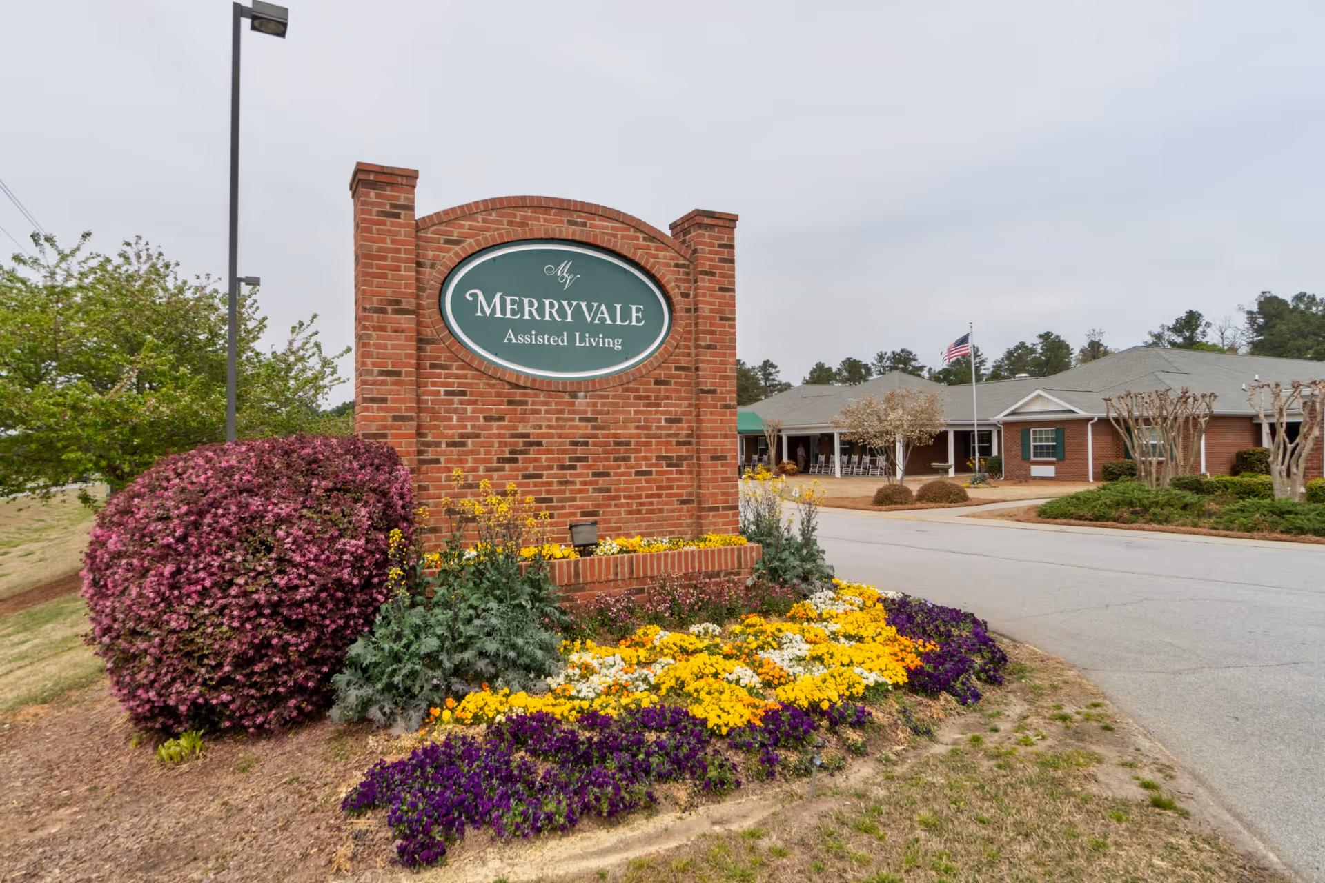 Brick sign for Merryvale Assisted Living surrounded by colorful flowers and bushes, with a single-story building and an American flag in the background under a cloudy sky.