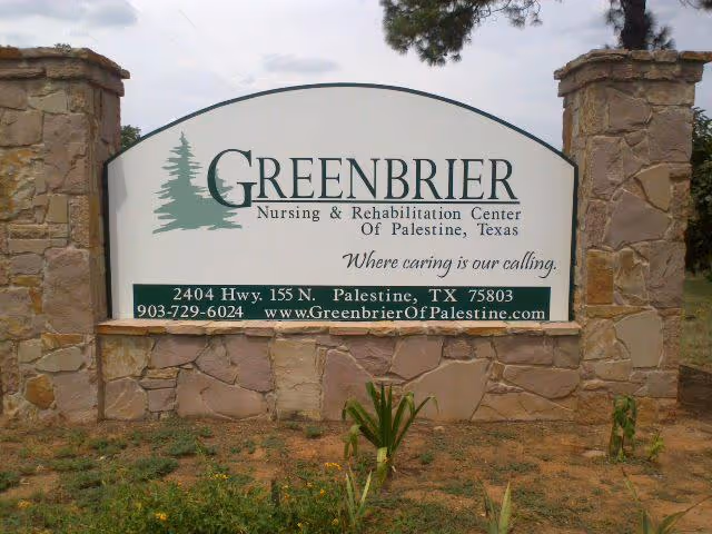 Stone signpost with a large white sign displaying the name Greenbrier Nursing & Rehabilitation Center of Palestine, Texas, along with the slogan 'Where caring is our calling.' The sign also includes the address 2404 Hwy. 155 N. Palestine, TX 75803, phone number 903-729-6024, and website www.GreenbrierOfPalestine.com. The sign is set outdoors with some plants and trees visible around it.