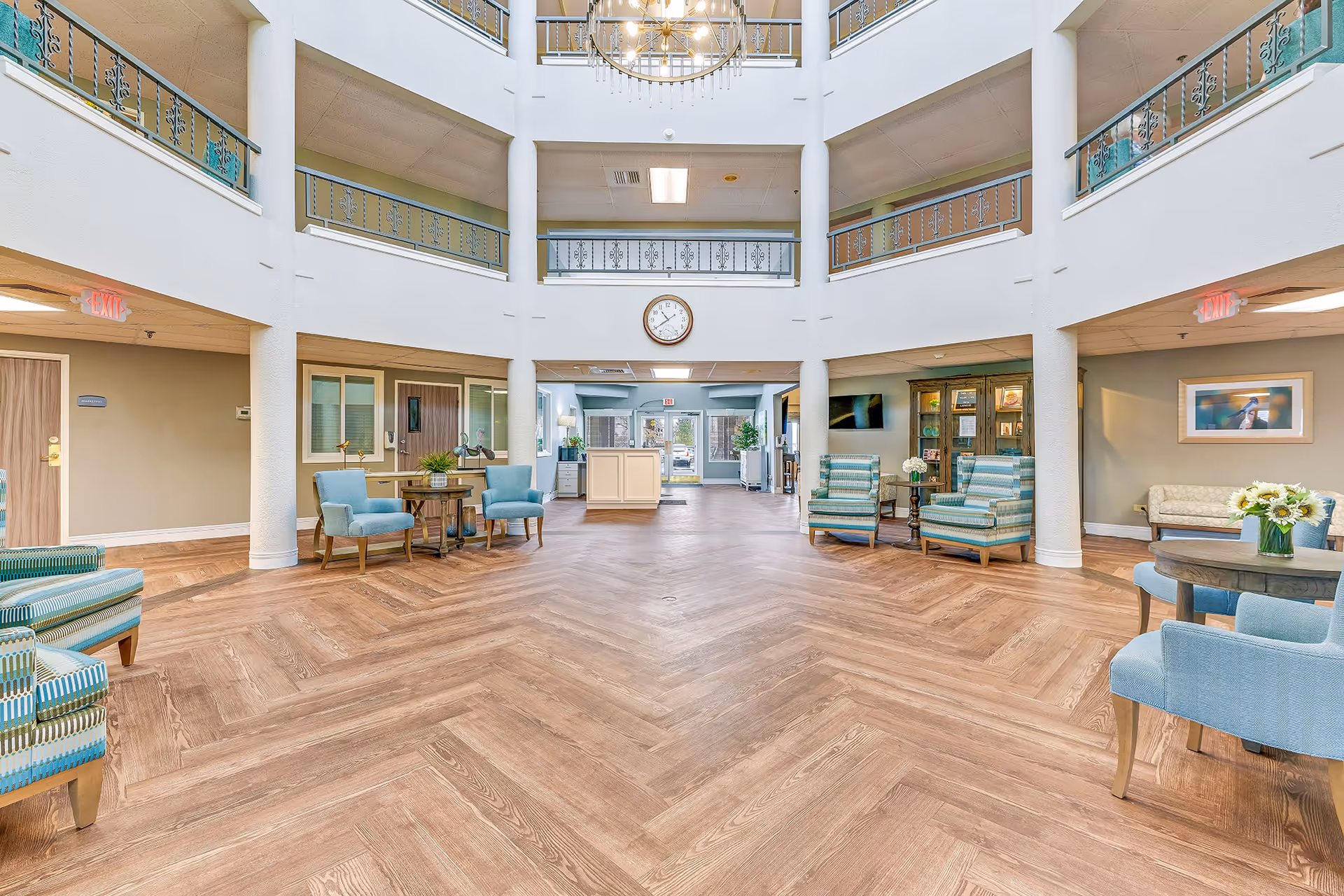 Spacious and bright senior living facility lobby with high ceilings and a large chandelier. The room features light wood herringbone flooring, multiple seating areas with blue and striped armchairs, small tables with plants and flowers, and a clock mounted on the far wall above a reception desk. The upper floors with decorative railings are visible, and exit signs are illuminated on both sides of the room.