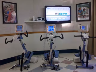 A small exercise room with three stationary exercise bikes facing a wall-mounted TV screen displaying the Wii Sports game menu. The room has light-colored walls with a wooden chair rail and two framed pictures hanging on the walls. The floor has a checkered pattern with light and dark tiles.