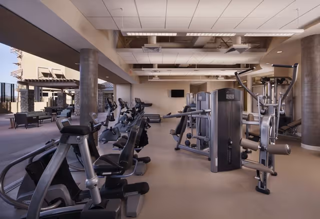 Indoor gym area with various exercise machines including stationary bikes and weight machines, adjacent to an outdoor patio seating area with tables and chairs under a pergola.