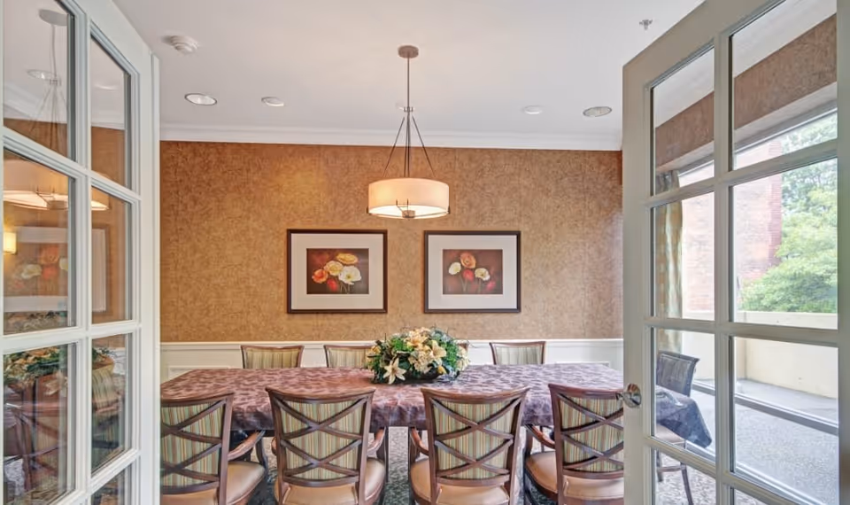 A dining room with a long table covered by a patterned tablecloth and surrounded by eight chairs with striped upholstery. A floral centerpiece is placed in the middle of the table. The walls are decorated with two framed floral paintings, and a modern pendant light hangs from the ceiling. French doors with glass panes open into the room, and a window shows an outdoor area with greenery.