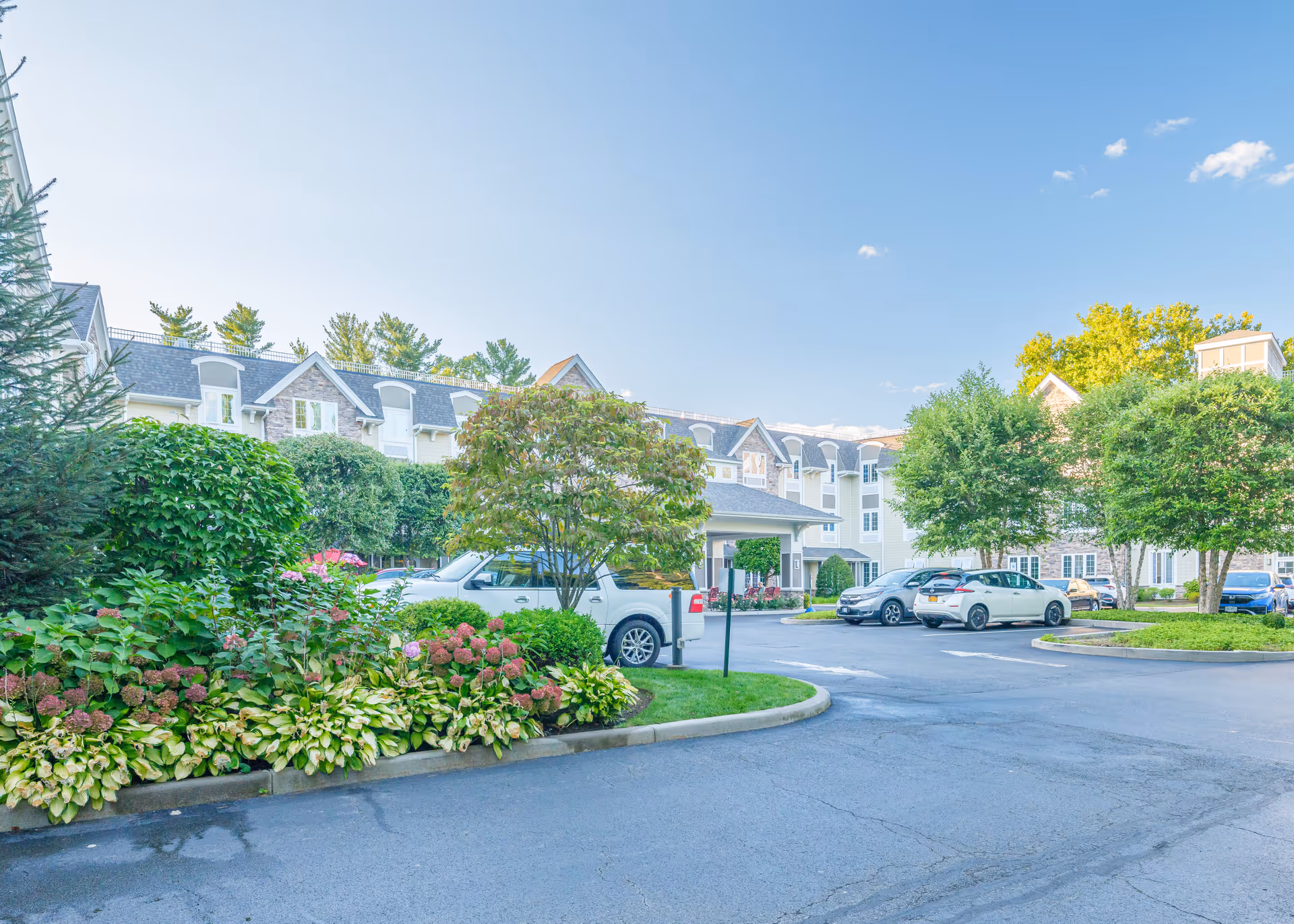 Front entrance and parking area of a senior living facility with landscaped flowerbeds, trees, and parked cars under a blue sky.