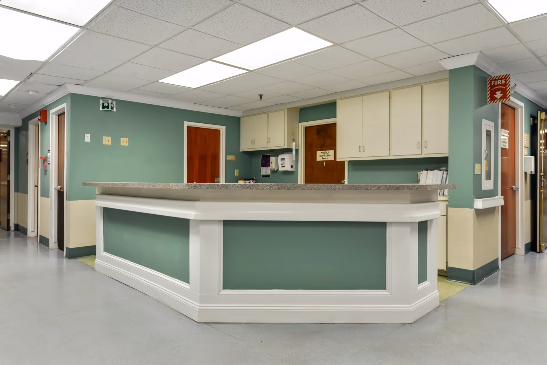 Reception or nurse station area in a healthcare facility with a green and white counter, green walls, wooden doors, overhead cabinets, and a fire alarm sign.
