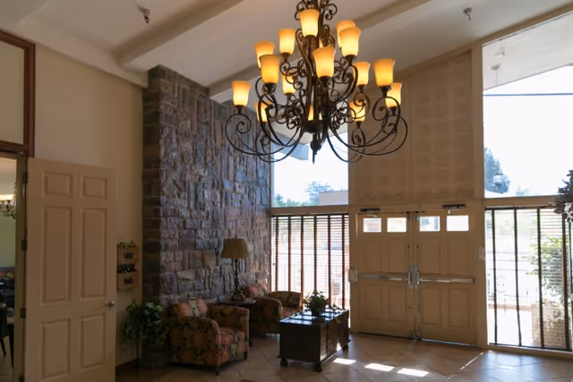 Interior view of a nursing facility lobby with a large ornate chandelier hanging from the ceiling, a stone accent wall, two patterned armchairs, a small table with a lamp, and double doors with windows letting in natural light.