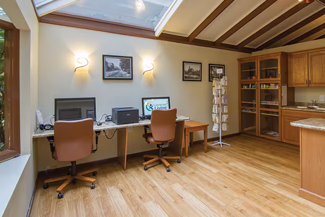 Bright common room featuring two computer workstations with rolling chairs, shelving and a small kitchenette under skylights.