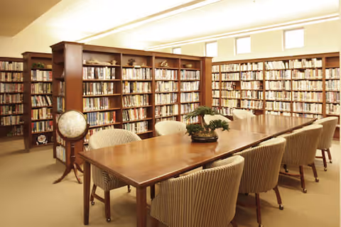 A well-lit library room with wooden bookshelves filled with books lining the walls. In the center, there is a long wooden table surrounded by cushioned chairs with striped upholstery. A small bonsai tree is placed on the table as decoration.