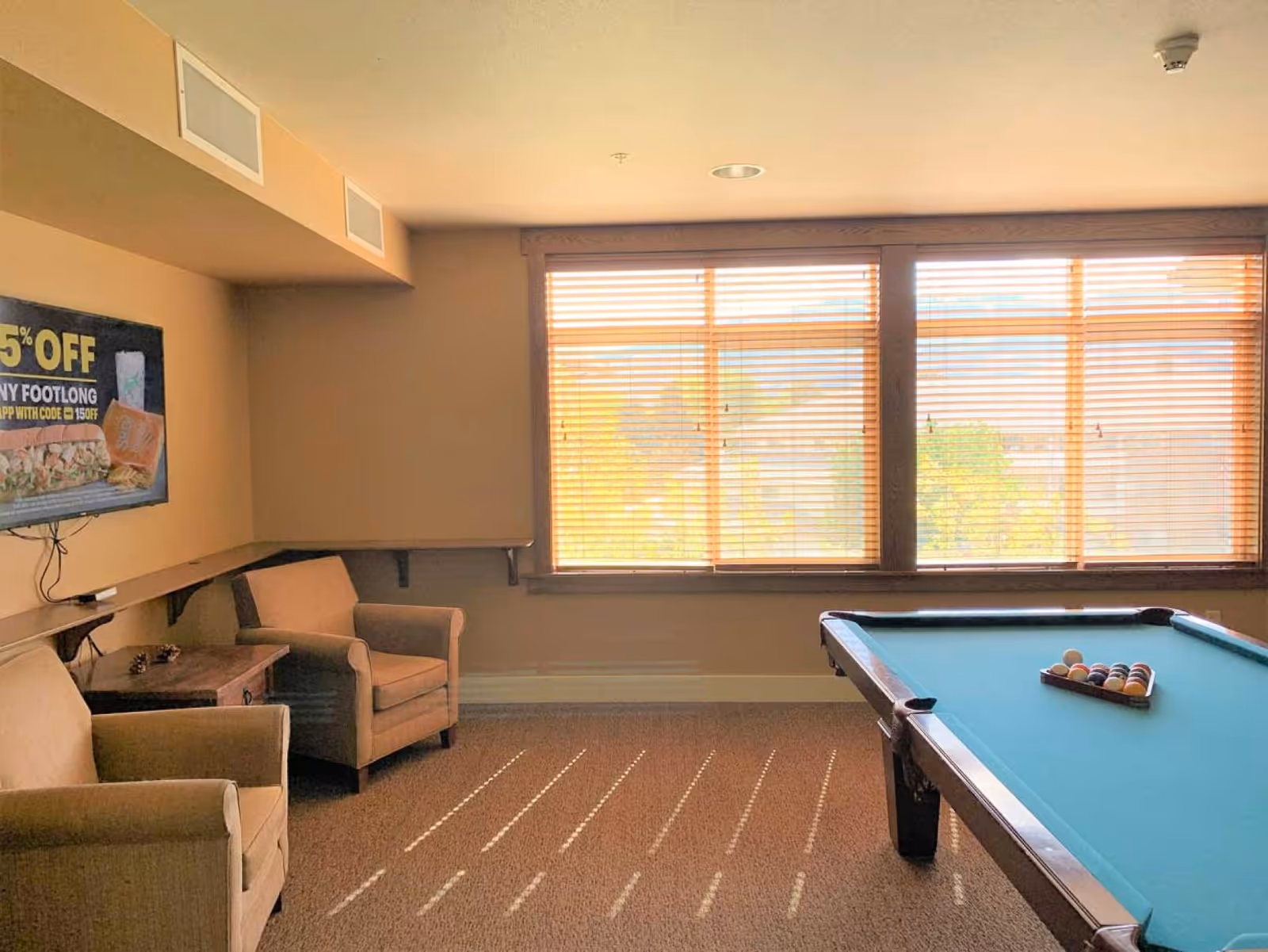 A cozy room with a pool table set up for a game, two beige armchairs, a small wooden side table, and a wall-mounted TV displaying a 15% off promotion. Large windows with wooden blinds allow natural light to fill the room.
