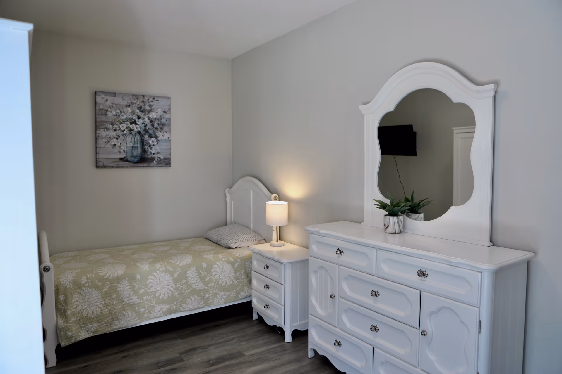 Small bedroom featuring a single bed with a patterned quilt, a nightstand with a lamp, and a white dresser topped by a mirror and plant.