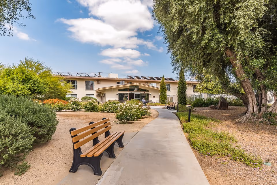 A paved walkway leading to the entrance of a two-story building with benches along the path, surrounded by bushes, trees, and landscaped garden areas under a partly cloudy sky.