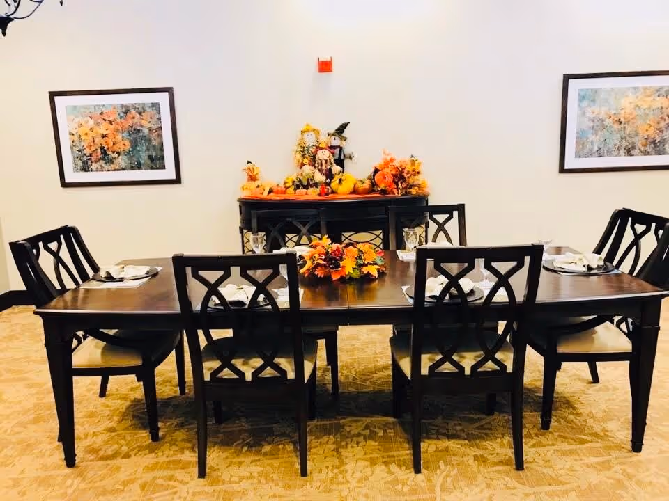 A dining room with a dark wooden table set for six people with white napkins and glasses. The table has a centerpiece of autumn-themed flowers and decorations. Behind the table is a sideboard decorated with fall-themed items including pumpkins and scarecrows. Two framed floral paintings hang on the light-colored walls.