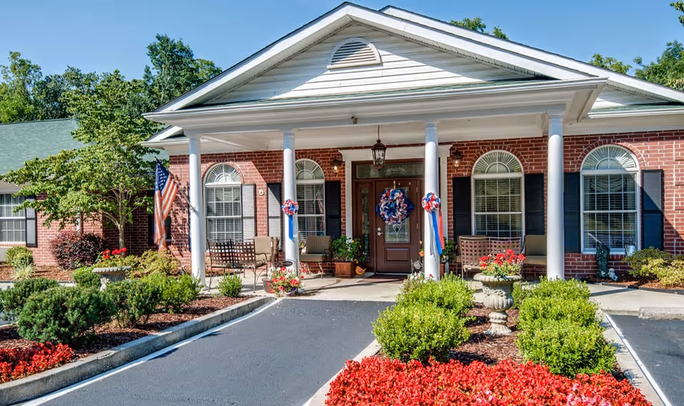 Front exterior view of a single-story brick building with white columns supporting a porch roof. The entrance door is decorated with a patriotic wreath, and there are additional red, white, and blue decorations on the columns. The building is surrounded by well-maintained landscaping including green shrubs, red flowers, and a small American flag on the left side. The sky is clear and blue.