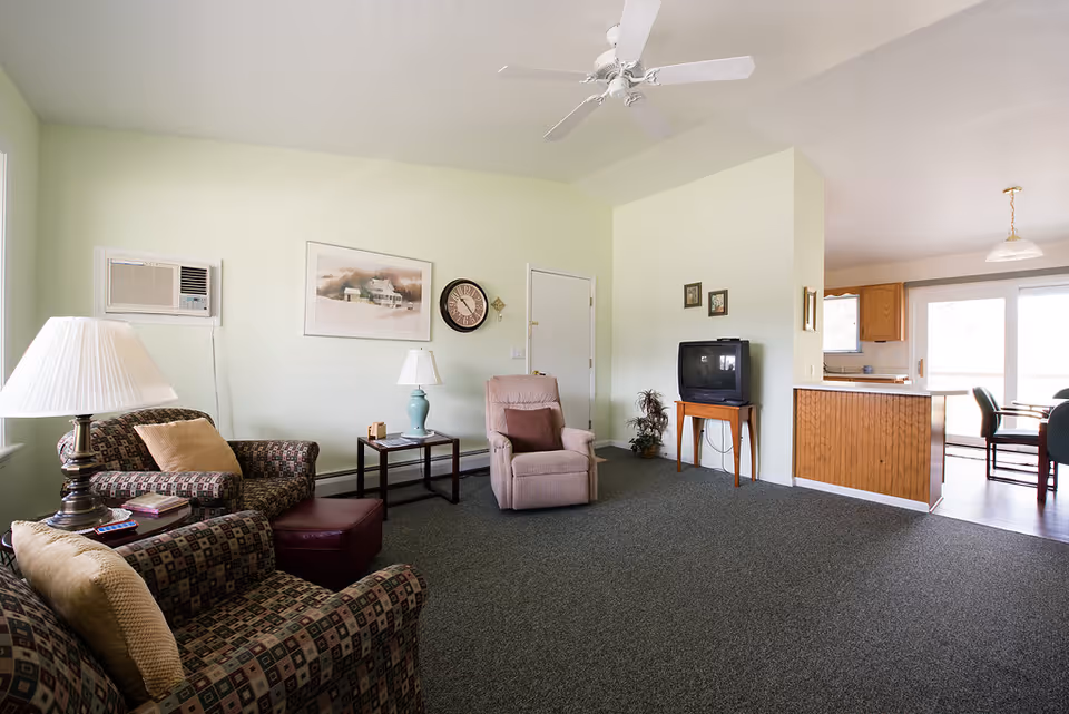 Spacious living room with patterned armchairs, a recliner, small tables and lamps, a TV on a stand, ceiling fan, and an open view into the kitchen/dining area.