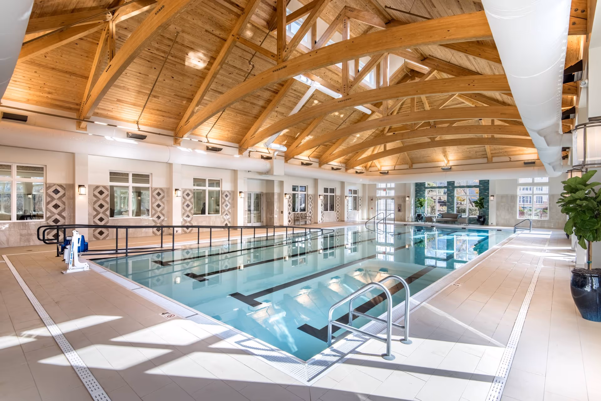 Indoor swimming pool with clear water, metal handrails, and a ramp for accessibility. The pool area has a high wooden ceiling with exposed beams and large windows letting in natural light. There are potted plants and seating areas near the windows.