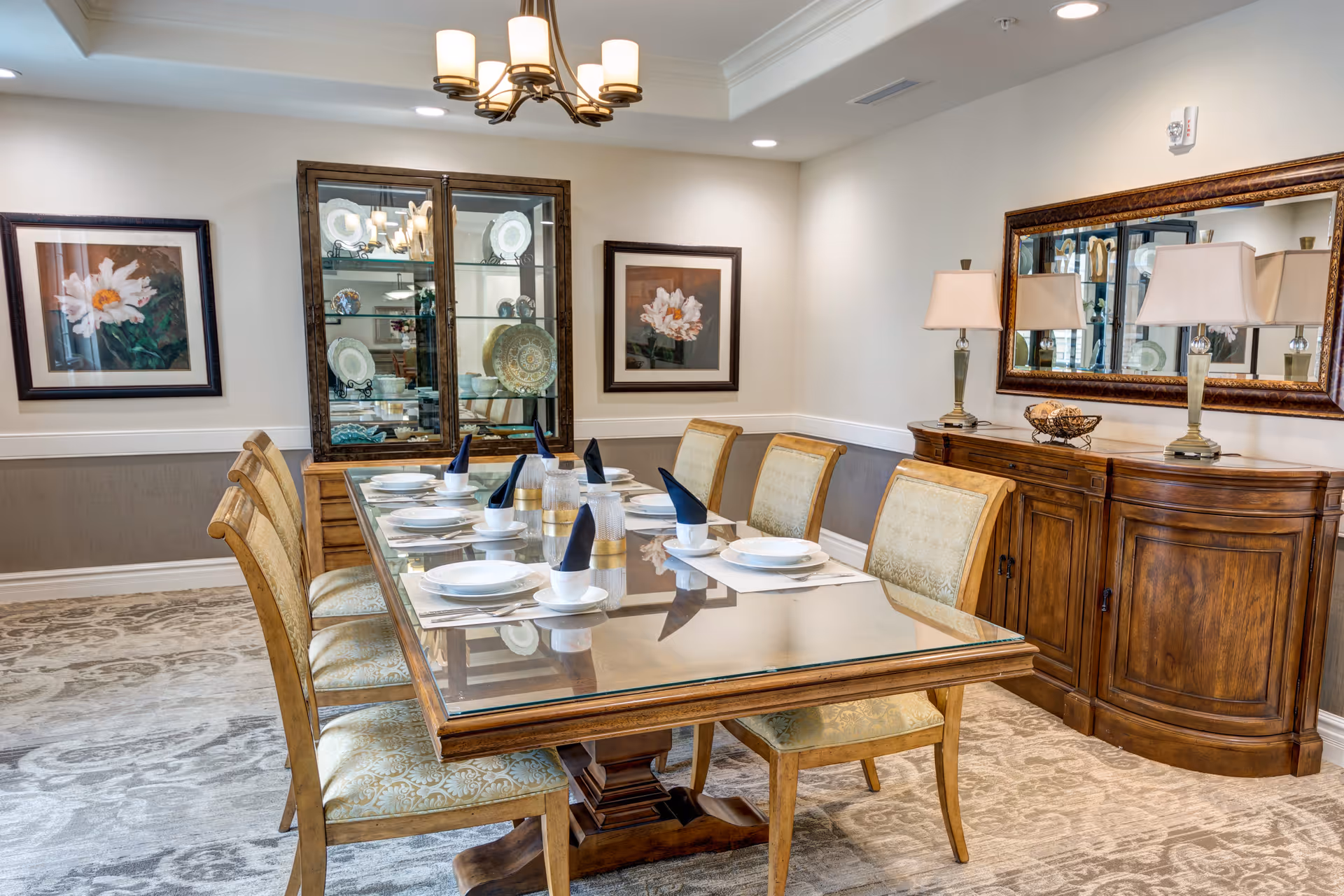 A formal dining room with a glass-top wooden table set for six people with white plates, bowls, cups, and black napkins. The room features a wooden sideboard with two lamps and a large mirror above it. There is a glass-front cabinet displaying decorative plates and two framed floral paintings on the walls. The floor is carpeted with a patterned rug.