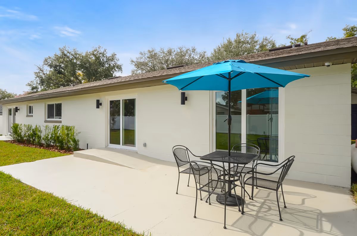 Outdoor patio area with a black metal table and four matching chairs under a large blue umbrella, adjacent to a single-story white building with sliding glass doors and windows, surrounded by green grass and trees.