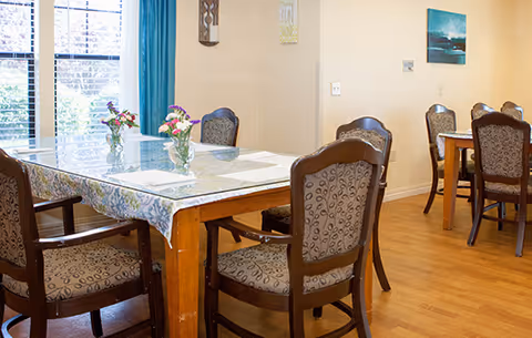 A dining area with wooden tables and cushioned chairs. Each table has a small vase with flowers and placemats. The room has wooden flooring, beige walls, and a window with blue curtains letting in natural light. There are framed pictures and wall decorations on the walls.