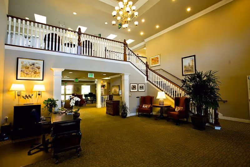 Spacious interior of a senior living facility with a large staircase featuring white railings and dark wooden handrails. The area is warmly lit with a chandelier and several lamps. There are two red armchairs with a small table and lamp between them near the staircase. A desk with a computer and plants is positioned on the left side. The walls are decorated with framed artwork, and there are large potted plants adding greenery to the space.