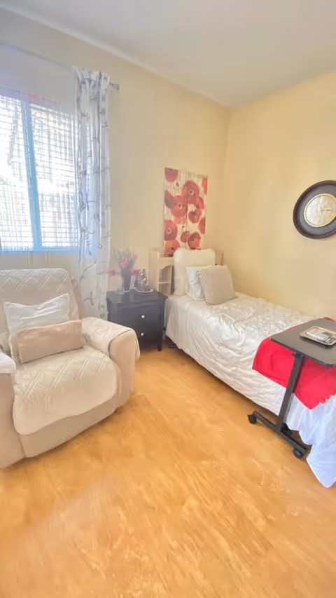A cozy bedroom with a single bed covered in white bedding and a red blanket at the foot. Next to the bed is a small black nightstand with decorative items and a vase of flowers. A comfortable beige armchair with cushions is positioned near a window with sheer white curtains. The walls are painted light yellow, adorned with a floral painting and a round wall clock.