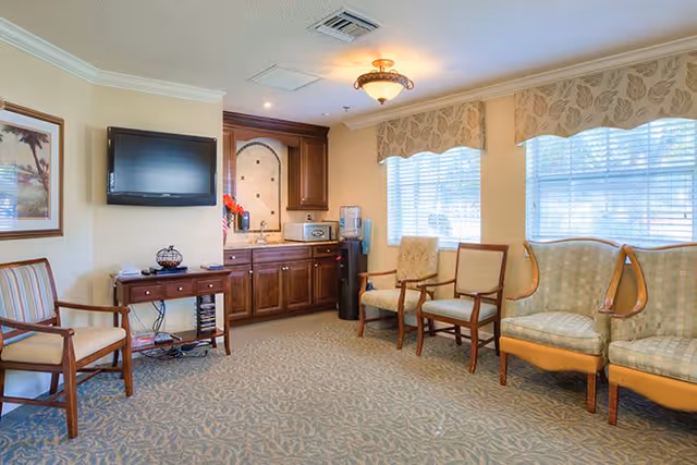 A well-lit common area with several wooden chairs with cushions arranged along the walls. There is a small wooden table with a decorative item and a flat-screen TV mounted above it. In the corner, there is a kitchenette area with dark wood cabinets, a microwave, a water cooler, and a small countertop. Large windows with patterned valances allow natural light to fill the room.
