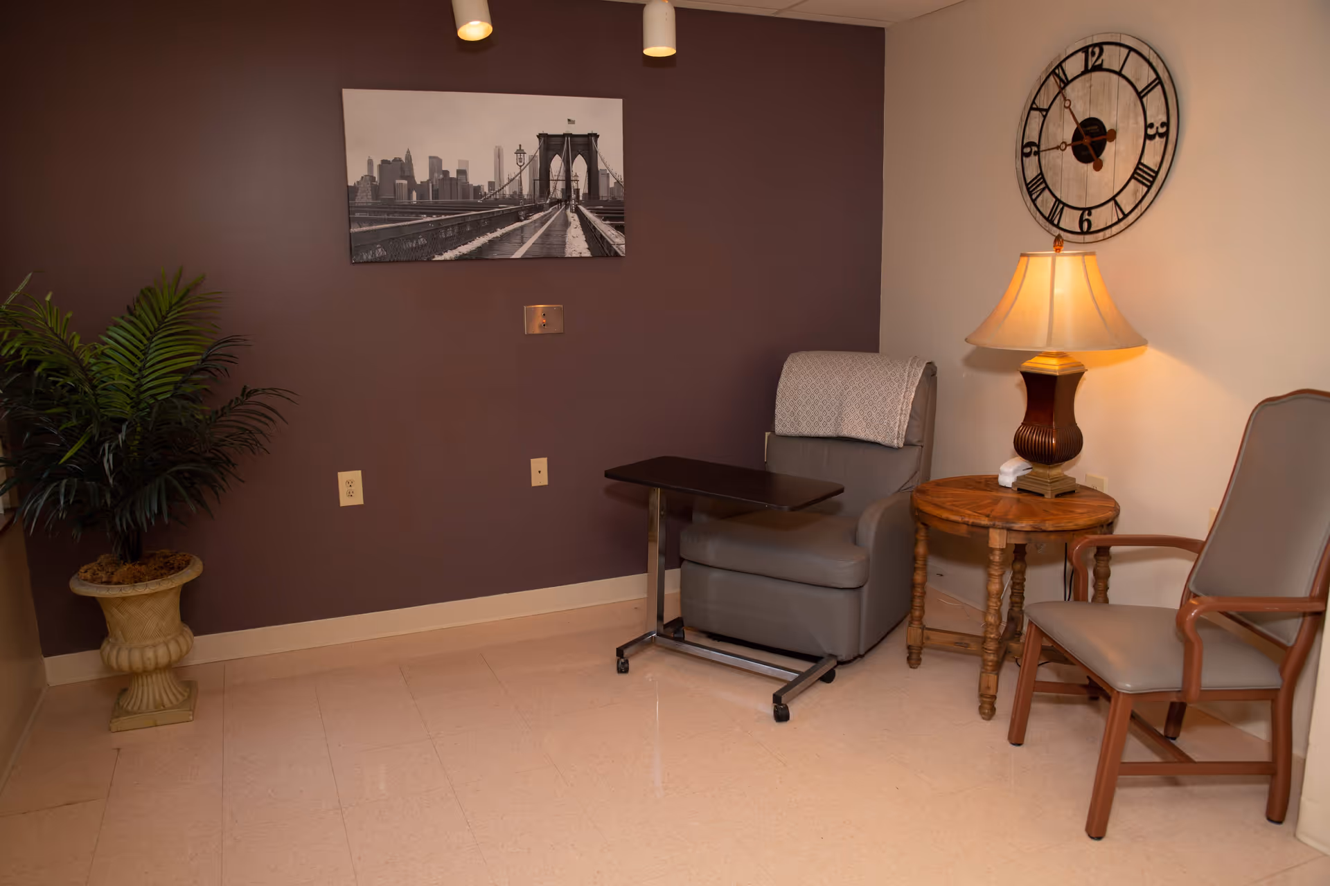 A cozy sitting area with a gray recliner chair featuring a small overbed table, a wooden side table with a lamp, and a wooden armchair. A large wall clock hangs on the white wall, and a black and white photo of the Brooklyn Bridge is mounted on a dark purple wall. A potted plant is placed in the corner.