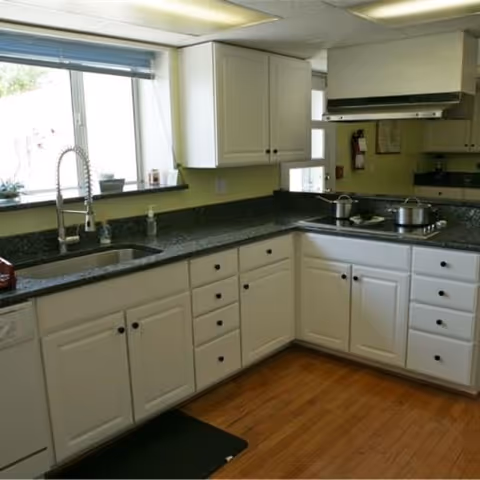 A clean kitchen with white cabinets, black granite countertops, a stainless steel sink with a modern faucet, a stove with pots on it, and a window with blinds letting in natural light.