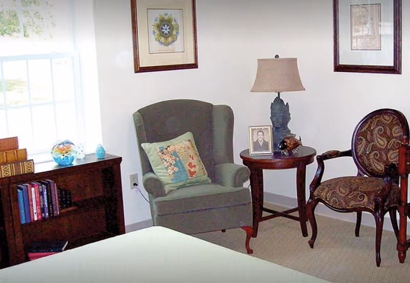 A cozy sitting area in a room featuring a green upholstered armchair with a decorative pillow, a round wooden side table with a lamp and framed photo, and a patterned wooden armchair. There is a wooden bookshelf with books and decorative items next to a window letting in natural light. Two framed pictures hang on the wall above the chairs.