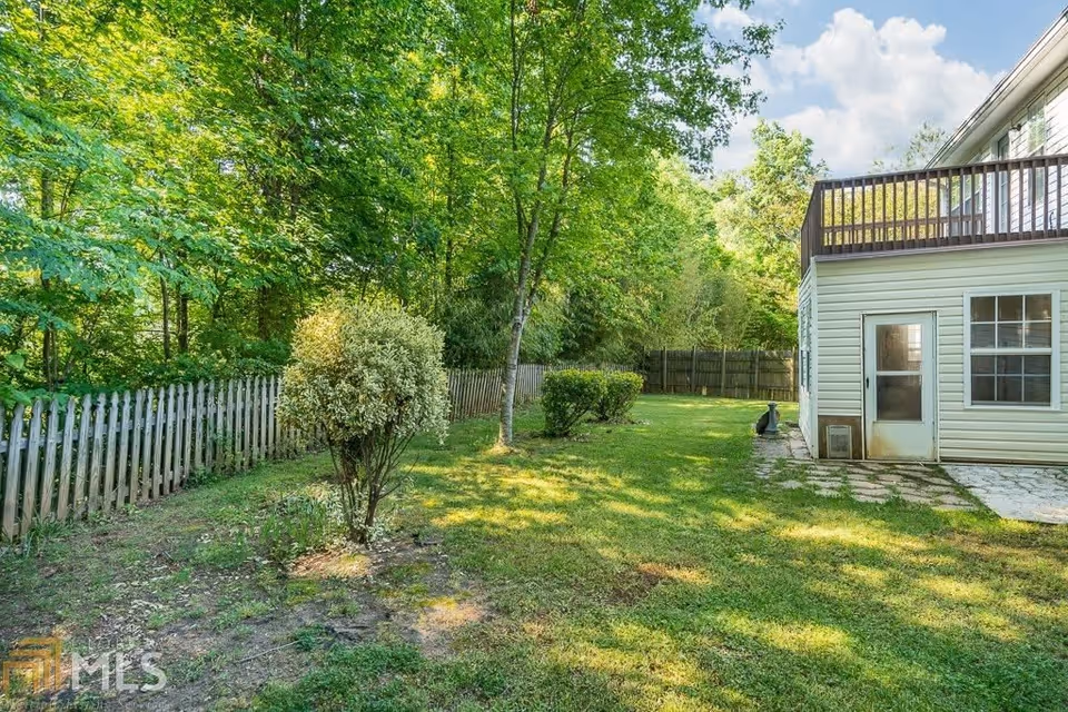 A backyard area with green grass, several bushes, and trees along a wooden picket fence. On the right side, there is a white two-story building with a door and windows, and a wooden balcony on the upper floor. The sky is partly cloudy with blue patches.