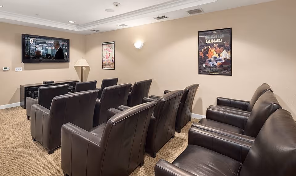 A small theater-style room with two rows of black leather chairs facing a wall-mounted flat screen TV. The walls are beige with two framed movie posters, one for Casablanca. The room is carpeted and has a floor lamp in the corner.