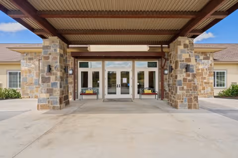 Entrance of a building with a covered driveway supported by stone pillars. The building has double glass doors with benches on either side, and windows on the walls. The sky is clear with a few clouds.