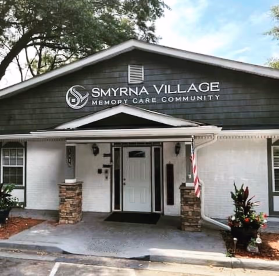 Front exterior view of Smyrna Village Memory Care Community building with a white door entrance, stone pillars, an American flag, and surrounding greenery.