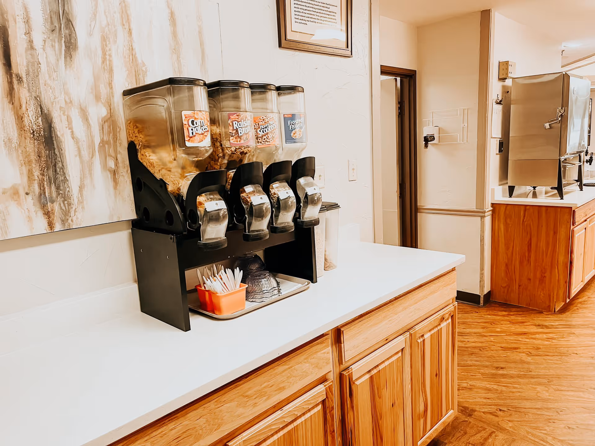 A countertop with a cereal dispenser containing Corn Flakes, Raisin Bran, Honey Nut Cheerios, and Frosted Flakes. Below the dispenser are small bowls and spoons. The area has wooden cabinets and a hardwood floor, with a beverage dispenser visible in the background.