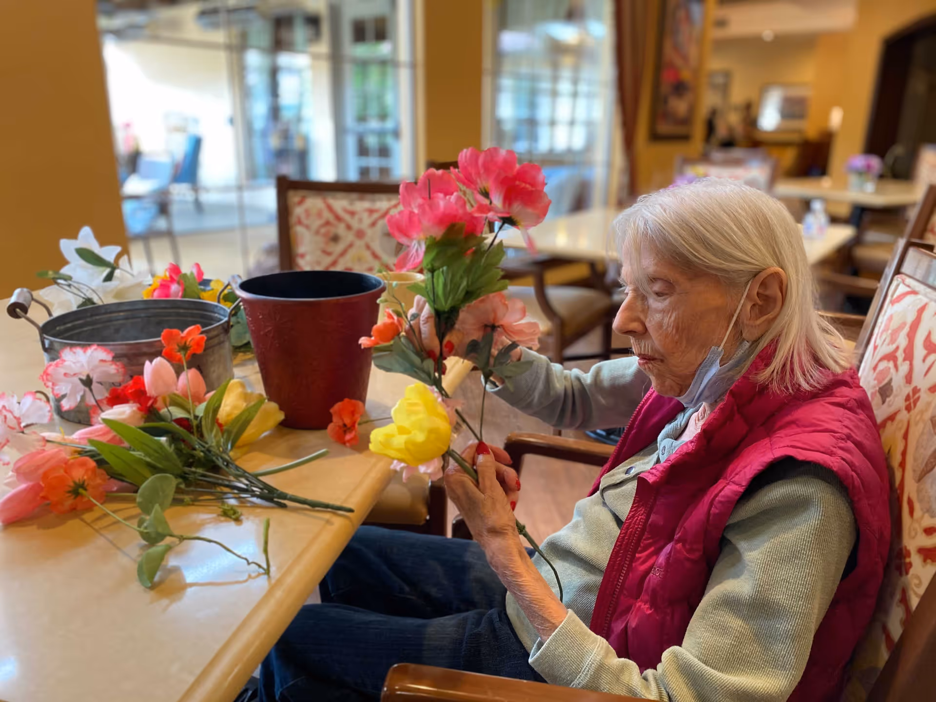 An elderly woman with white hair wearing a red vest and a face mask under her chin is sitting at a table arranging colorful artificial flowers in a bright, cozy room with patterned chairs and large windows.