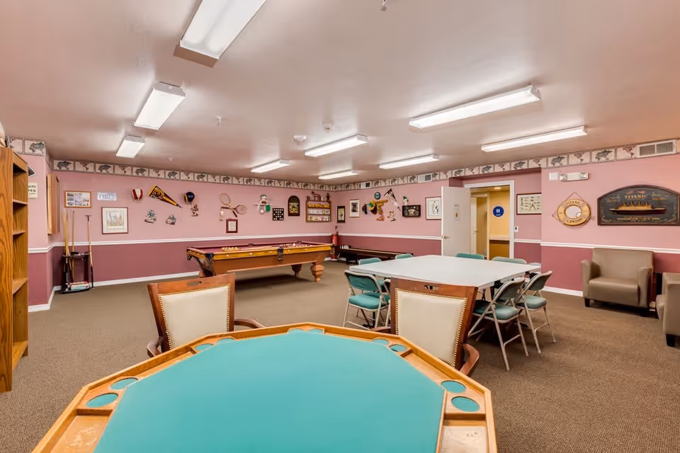 A recreational room in a retirement community featuring a green felt card table in the foreground, a pool table in the middle, and a rectangular table with chairs. The walls are decorated with various framed pictures, sports memorabilia, and nautical-themed items. The room has pink walls with a white chair rail and beige carpet, with fluorescent ceiling lights providing illumination.