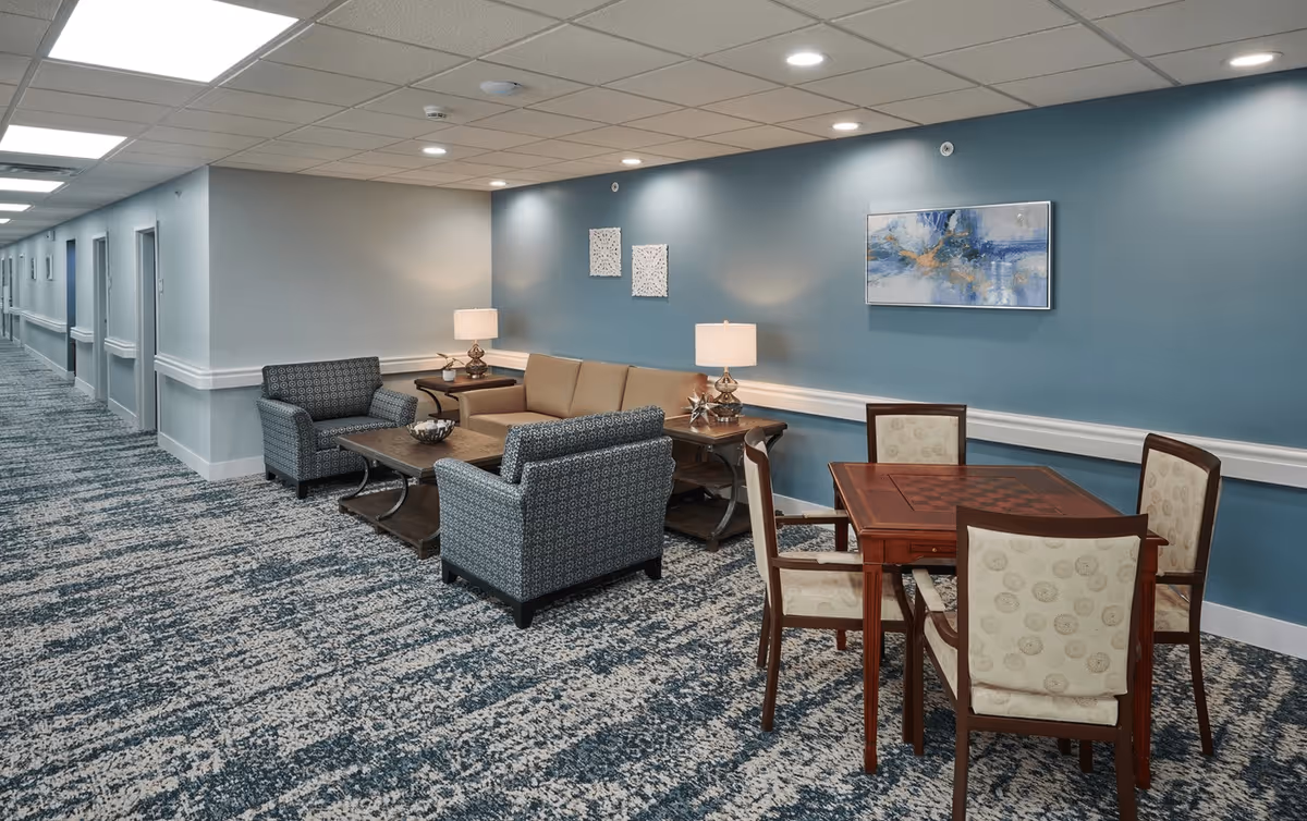 A senior living facility common area with a blue accent wall, two patterned armchairs, a beige sofa, two side tables with lamps, a wooden table with four chairs, and a hallway with multiple doors.