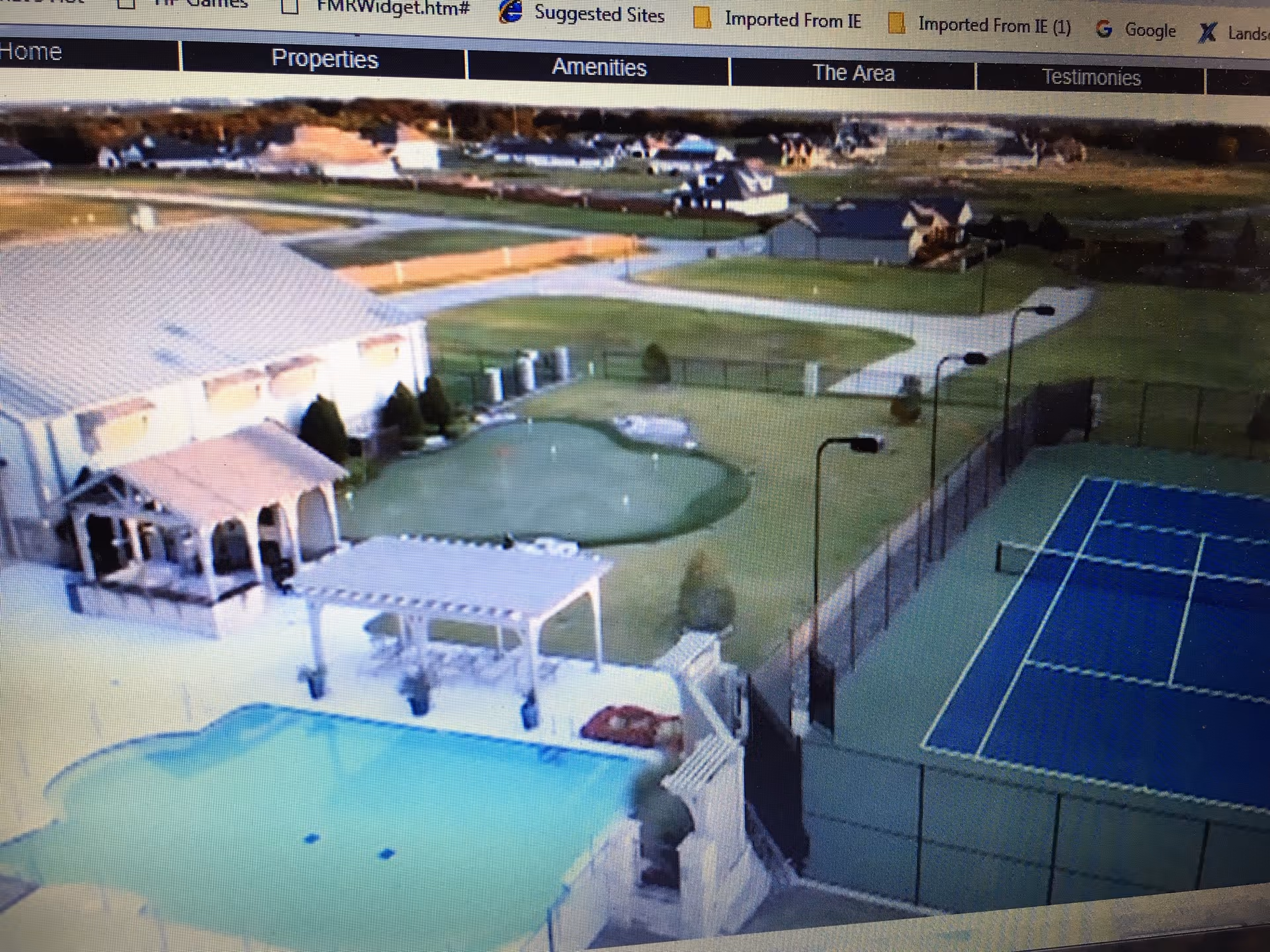 Aerial view of an outdoor recreational area at a senior living facility featuring a swimming pool, a covered patio with seating, a small putting green, and a fenced tennis court. Surrounding the area are pathways, grassy fields, and residential houses in the background.
