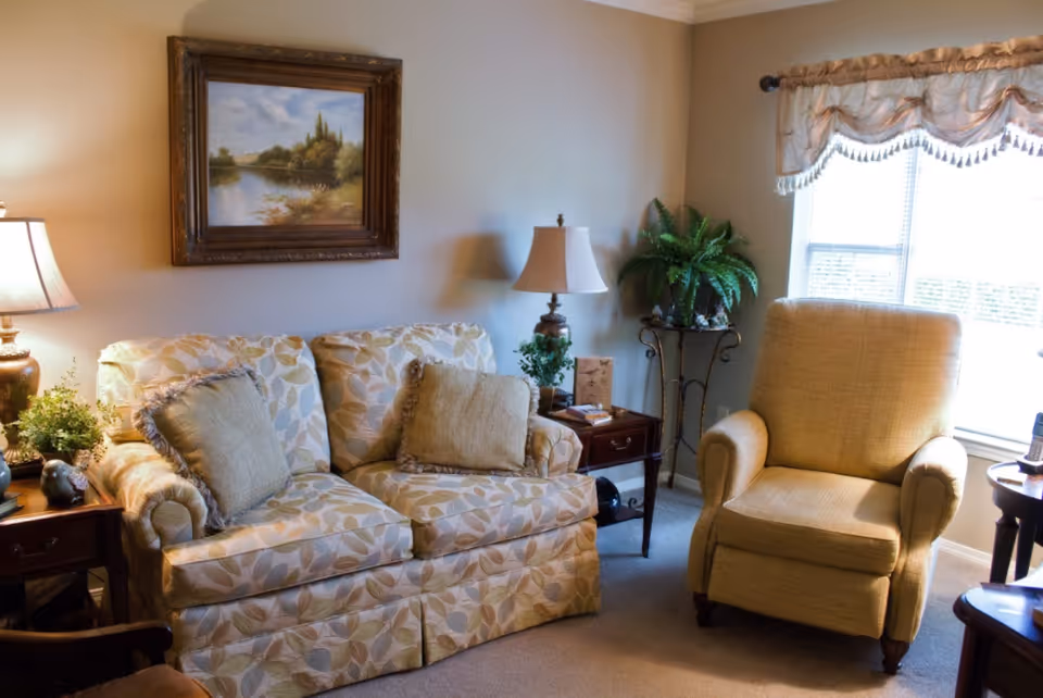 A cozy living room with a patterned loveseat with two cushions, a beige armchair, two wooden side tables with lamps and plants, a framed landscape painting on the wall, and a window with decorative valance curtains letting in natural light.