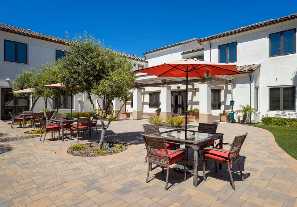 Sunny outdoor courtyard with patio tables and red umbrellas in front of a two-story white building.