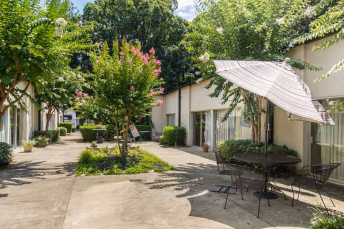 Outdoor courtyard area with a concrete walkway, surrounded by single-story buildings with large windows. There are trees and shrubs planted along the walkway, and a round metal table with four chairs and a large striped umbrella providing shade.