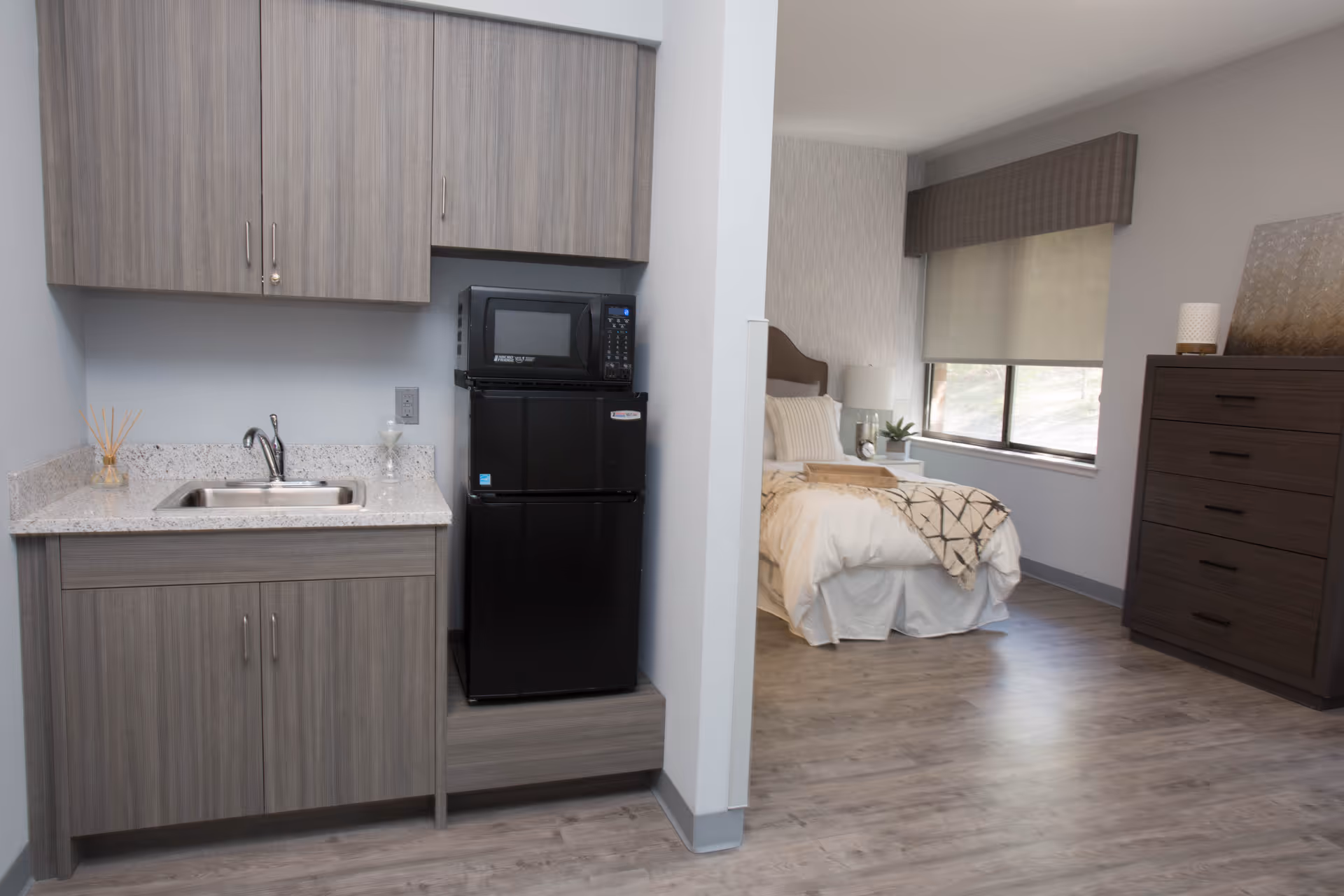 A small kitchenette area with a sink, countertop, upper and lower cabinets, a black microwave, and a black mini refrigerator. Adjacent to the kitchenette is a bedroom with a bed, bedside table with a lamp, a window with a shade, and a dark wooden dresser.