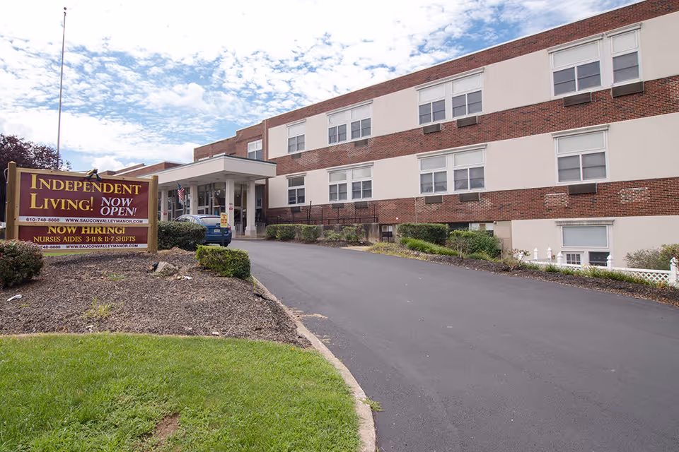 Exterior view of Saucon Valley Manor, a multi-story brick and beige building with several windows. A driveway leads to the main entrance with a covered drop-off area. A large sign near the entrance reads 'Independent Living! Now Open!' and advertises hiring for nurses and aides. The sky is partly cloudy.