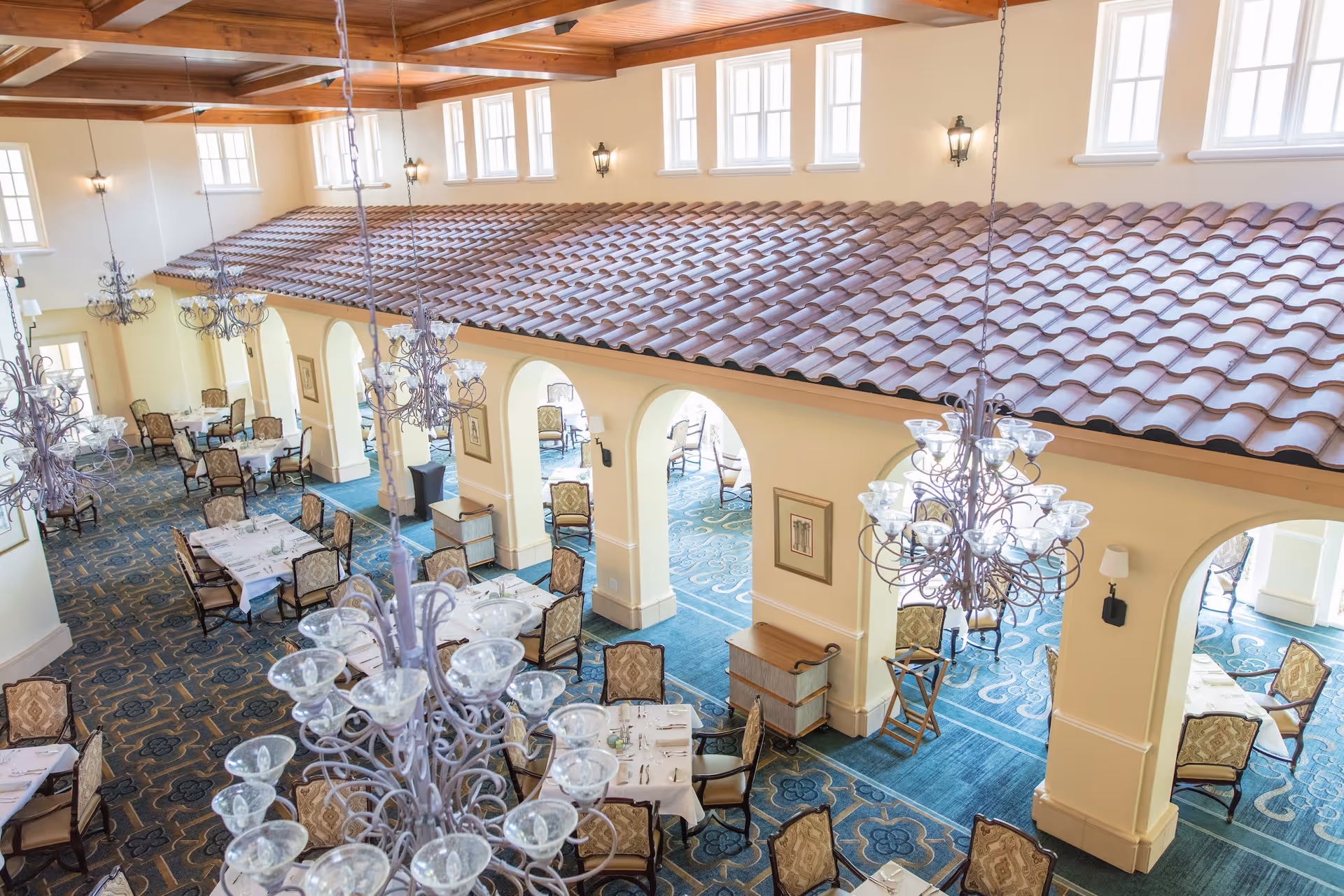 A spacious dining room with multiple tables set with white tablecloths and chairs arranged neatly. The room features a high ceiling with wooden beams, large chandeliers, arched doorways, and multiple windows allowing natural light to fill the space. The floor is covered with a patterned blue carpet.