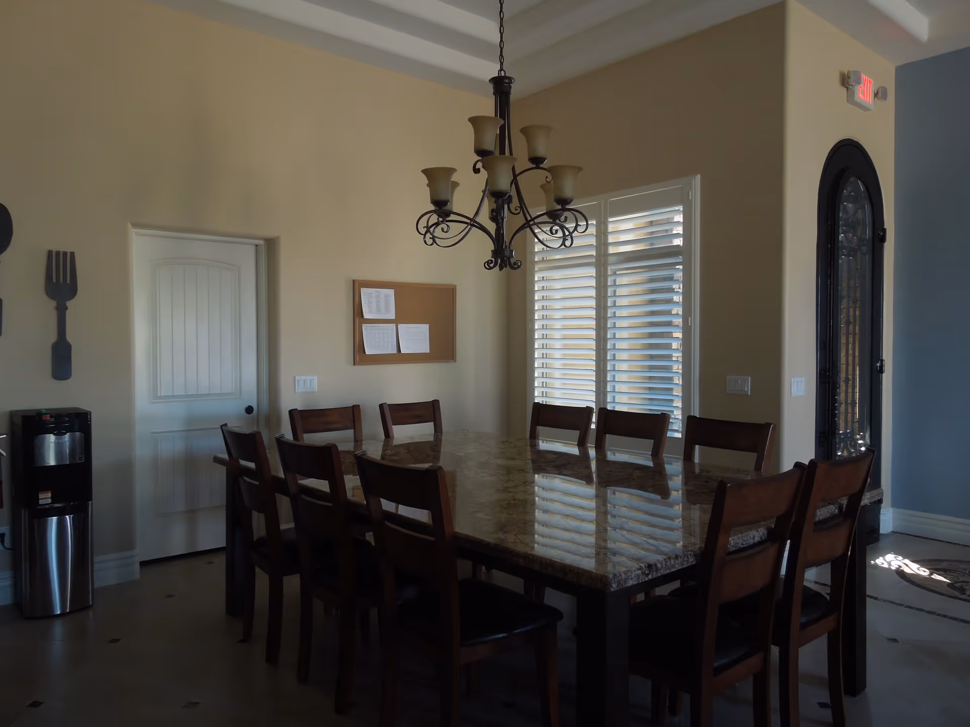 A dining room with a large rectangular marble table surrounded by ten wooden chairs. A chandelier with multiple lights hangs above the table. There is a window with white shutters on the right wall and a door on the left wall. A water dispenser is placed near the door, and decorative oversized fork and spoon wall hangings are visible on the left side.